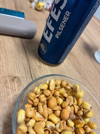 A high-angle shot captures a scene on a wooden table. A blue can of Efes Pilsener beer is positioned to the left, partially out of focus. Adjacent to the can is a gray, rectangular device, possibly a vape pen. A clear glass bowl filled with a variety of roasted nuts dominates the right side of the image. The lighting suggests an indoor setting. There is no visible indication of the image being taken in Dulovo, Bulgaria.