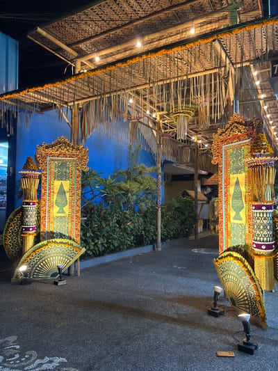 The image depicts an outdoor setting in Pecatu, Indonesia, at night. There is a structure overhead, constructed from bamboo and woven palm leaves, and decorated with strings of orange flowers and small lights. Along a paved walkway are two decorative displays, featuring framed artwork with ornate gold borders and images of what appears to be traditional vases. These displays also incorporate parasols, floral arrangements, and patterned fabrics. Behind the displays, there are bushes and a blue wall. There are also wall-mounted spotlights illuminating the scene.
