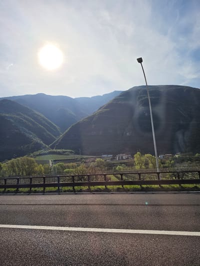 The image shows a view of a landscape in Avio, Italy, taken from a bridge or elevated roadway. Mountains rise in the background, partially obscured by haze. The sun is visible in the sky, causing glare. A lamppost stands to the left of the mountains. Below the mountains, there's a visible landscape with trees, vineyards, and some buildings that appear to be houses. A metal railing runs along the side of the roadway, separating it from the view. The roadway itself is made of asphalt with a white dividing line.