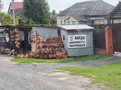 The image captures a roadside market stall in Novye Vyselki, Russia, specializing in woven baskets. In the foreground, a gravel driveway is bordered by patches of green grass. A woman in a black shirt and dark skirt is seated on a metal chair, facing away from the camera, seemingly tending to the stall. To her left, shelves display various items, including books or magazines.
The main focus is the market stall itself, constructed with corrugated metal and wood. It is heavily laden with a diverse collection of wicker baskets of different sizes and shapes. Some are displayed hanging from the stall's structure, while others are neatly arranged on shelves and on the ground. Beyond the stall, a rustic building with a gray, corrugated metal roof is visible, and further back, a traditional Russian house with a red roof and wooden siding stands. A brick pillar and a dark brown fence are also present. A sign on the corrugated metal structure reads "НАРДЫ ШАХМАТЫ МАТРЕШКИ" in Russian, indicating that games such as Nard, Chess, and Matryoshka dolls are also sold. The overall scene suggests a casual, rural marketplace.