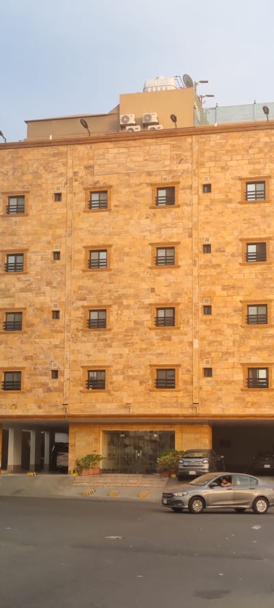 The image shows a five-story building in Jeddah, Saudi Arabia, constructed with yellow stone cladding. The building has evenly spaced rectangular windows with horizontal bars. On the roof, there are air conditioning units and a water tank. At the base of the building, there is a glass entrance and a parking area. Several cars are visible; one is a silver sedan driving on the road in front of the building, with a person visible in the driver's seat. Potted plants flank the entrance, and the sky is light blue.
