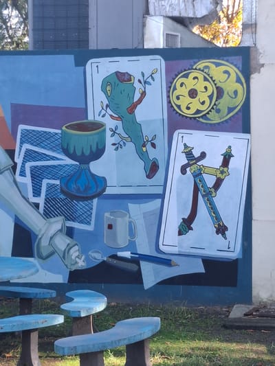 The image shows a mural in Villa Domínico, Argentina. The mural depicts several objects and playing cards in a surreal composition. In the upper left corner are two stylized yellow gears. To the right, are two playing cards with swords. The top card has two swords crossing each other. Below this, is another playing card with a green figure that resembles a plant-like monster. Below this card, are the rim of a goblet and a stack of playing cards. Next to them are a white coffee mug, a pencil and a spoon on a sheet of paper. Finally, the base of a chess king piece is featured along the bottom edge of the mural. The mural appears to be painted on a wall or surface next to an outdoor area. Part of a building is visible in the upper left corner and grass is seen in the lower right corner. A blue bench with multiple curved wooden slats is to the right of the mural.