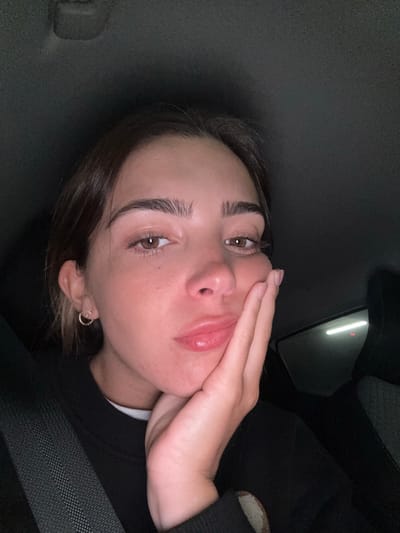 A young woman with light brown hair and eyes poses in a car. Her right hand is gently placed against her cheek. She is wearing a black sweater and a seatbelt is visible diagonally across her body. The car's interior is dark, with a rectangular light source visible through the window. The setting is most likely not in Senhora da Hora, Portugal, as there are no identifying features in the image that would suggest that location.