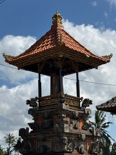 The image shows a close-up of the top of a Balinese temple structure against a blue sky with scattered clouds. The temple has a tiered roof covered with orange tiles and intricate golden ornamentation along the eaves and on the pinnacle, which features a crown-like finial. The structure below the roof is open, supported by dark pillars, and features decorative golden panels with carved figures and a stone base with detailed carvings of mythical creatures. Palm trees are visible in the background on the left and right sides of the frame, suggesting a tropical environment. The scene is located in Tegallalang, Indonesia. There are no people or animals visible in the image. The lighting indicates it is daytime.