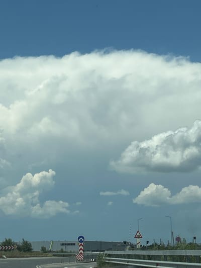 The image captures a daylight scene in Tsaratsovo, Bulgaria, depicting an industrial landscape under a partly cloudy sky. The upper portion of the frame features a bright blue sky, with a substantial, dense bank of white cumulus clouds extending across the top-middle, and smaller, more dispersed white and grey clouds scattered below and to the right.
In the foreground, a curved section of asphalt road is visible, suggesting a roundabout or an intersection. A white metal guardrail runs along the right edge of this road segment. To the left of the road, green foliage, including trees and shrubs, lines the roadside.
In the mid-ground, a prominent multi-colored road sign is erected on a red and white striped pole. The sign is a blue circle containing two white arrows pointing downwards in opposite directions, typically indicating a traffic island or advising to pass on either side. Further back, a large, rectangular industrial building, grey in color, stretches across the horizon. The name "LIEBHERR" is clearly visible on the side of this building. Adjacent to the building, a piece of green industrial machinery, possibly a crane or lift, is partially visible.
To the right of the road, another triangular red warning sign, featuring a symbol for an uneven road or speed humps, stands on a pole. Two modern streetlights with tall, dark poles are positioned further to the right, beyond which more green vegetation and indistinct industrial structures are visible in the distance. No individuals are present in the image.