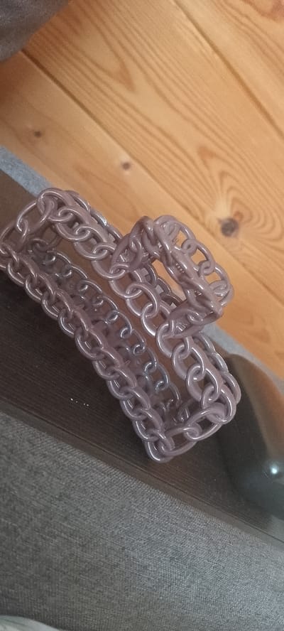 A chain lies on a wooden surface next to a gray fabric surface. The chain is made of interconnected links. The wooden surface appears to be made of planks. The gray fabric surface is textured. The image appears to be taken indoors, possibly in a residential setting. There is no visible indication of the city of Terzi, Russia, or any other specific location.