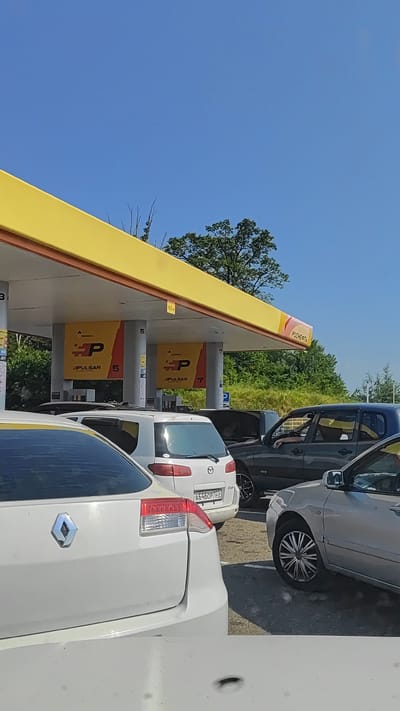 A line of cars is parked at a gas station in Razdolnoye, Russia. The gas station canopy is yellow with red trim and has signs with the logo "HP PULSAR." The sky is clear blue. The cars are a white Renault, a white Mazda, and a dark gray SUV. The white Renault is in the foreground, and the white Mazda is behind it. The dark gray SUV is behind the white Mazda. The gas station appears to be open, as the pumps are visible and the canopy is lit. The image captures a typical scene at a gas station.