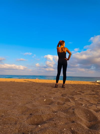 An overhead shot captures a person standing on a sandy beach in Nahariyya, Israel, facing the ocean. The person, with tattoos visible on their back and arms, is wearing a black tank top and black leggings. They are standing with their right hand on their hip and their left hand behind their head, looking out at the water. The ocean stretches out to the horizon, meeting a bright blue sky dotted with white clouds. The sand is a warm, golden brown, with ripples and scattered debris indicating a natural beach environment. A shadow stretches across the sand from the person, cast by the warm light of the sun. The scene suggests a moment of contemplation or rest by the sea.