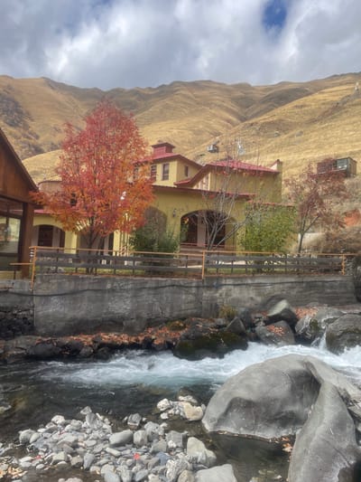 This image captures a scenic view in Juta, Georgia, presenting a multi-story building complex situated alongside a river at the base of expansive mountains. The central structure is a light yellow building with multiple red-tiled roofs, featuring arched openings on its lower level and several windows on upper floors. A prominent deciduous tree with vibrant orange and red autumn foliage stands in front of this building, to its left. Part of a wooden building with large windows is visible on the far left. A wooden fence runs atop a grey concrete retaining wall, separating the elevated grounds of the buildings from the river. The river itself is fast-flowing, displaying white rapids and containing numerous large and small rocks. Scattered autumn leaves, primarily in red and orange hues, are visible on the riverbanks and within the water. In the background, large, treeless mountains covered in dry, golden-brown grass ascend steeply under a partially cloudy sky with patches of blue. A smaller, dark rectangular structure, possibly a cabin or container, is visible on the upper slope of the mountain to the right. No people are present in the scene.
