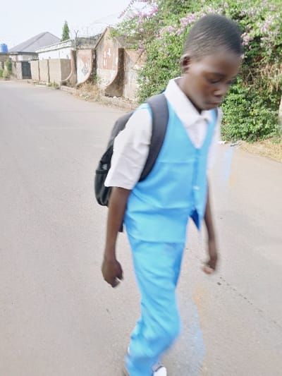 A young dark-skinned male, appearing to be a school-aged child, is depicted walking on a light-grey paved road in the city of Kwekwe, Zimbabwe. He wears a white collared shirt, a light blue sleeveless vest, and light blue trousers, with a dark-colored backpack on his shoulders and white shoes. His head is bowed, and his face exhibits motion blur, indicating movement. To the left of the road, several light-colored buildings with dark roofs are visible, including one with a dark gate, a tall green tree, and a blue water tank on a distant rooftop. To the right, a weathered concrete wall with decorative buttresses and sections of barbed wire runs parallel to the road, partially obscured by dense green foliage interspersed with pink-purple flowers. The sky is overcast.