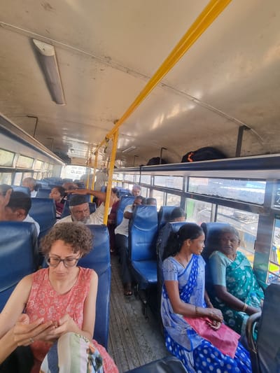 The image displays the interior of a public transit bus, observed from a forward-facing perspective, likely in Gorantla, India. The bus features rows of blue hard plastic seating, a worn off-white ceiling with exposed fluorescent light fixtures, and prominent yellow metal grab rails extending along its length. Overhead luggage racks are visible, one containing a black and orange bag. Blurred views through the windows reveal an exterior environment consistent with an urban or semi-urban setting, with some discernible text in Kannada script and English ("ELECTRICIAN WORK").

In the immediate foreground, a light-skinned woman with curly brown hair and glasses is seated, wearing a coral-patterned sleeveless top. She is looking down and manipulating a smartphone with both hands, wearing a ring on her left ring finger. Behind her, a bearded man wearing a dark skullcap and a striped shirt is seated, with his right arm extended in a distinct gesturing motion towards the right side of the bus. Further to the right, two women are seated; one younger woman in a blue patterned sari with gold earrings looks left with a slight smile, while an older woman beside her, with gray hair and a bindi, dressed in a teal-green patterned sari, looks directly forward. Multiple other passengers, predominantly adults in traditional Indian attire, are visible seated and standing throughout the bus. The metal floor features a textured, possibly corrugated, surface.