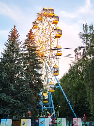 This is a vertical, eye-level, medium shot of a Ferris wheel in an amusement park during the daytime. The Ferris wheel is yellow with blue support beams. Several yellow cabins are visible, some with people inside. Tall evergreen trees are in the foreground on the left, with lush green deciduous trees behind them on the right. The sky is a soft blue with light clouds. The lighting suggests it is late afternoon or early evening, with warm sunlight illuminating the Ferris wheel and trees.

The scene is set in an amusement park, likely Yoshkar-Ola, Russia, as indicated by the Russian text on the posters at the bottom of the image. The posters, which are partly visible and feature the text "ПУТЕШЕСТВИЕ В ДЕТСТВО" (Journey into Childhood), suggest a family-friendly attraction. The overall mood is cheerful and nostalgic. In the lower portion of the image, there are signs and a fence, implying the entrance or perimeter of an attraction. Other amusement park rides or structures are partially visible behind the trees.
