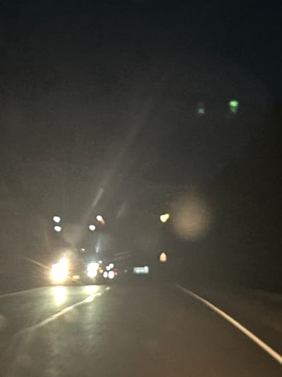 The image depicts a blurred, nighttime scene, likely captured from inside a vehicle. The primary subjects are the lights of other vehicles on what appears to be a road. In the distance, headlights and taillights create streaks and blobs of light against a dark background. There are also some green lights visible on the left side of the frame, which could be streetlights or indicators on a vehicle.
The setting is clearly outdoors at night. The blurred nature of the image suggests movement, possibly from the vehicle the photo was taken from. The overall atmosphere is dark and somewhat indistinct due to the lighting conditions and lack of sharp focus. There are no discernible people or specific objects beyond the vehicle lights.
The time of day is night. No specific weather conditions can be determined, though the haziness might suggest mist or rain, or it could simply be the result of poor image quality and low light. Location cues are absent beyond the context provided. No text is visible in the image.