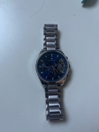 A single Tommy Hilfiger men's wristwatch with a silver-toned metal case and bracelet is positioned vertically on a flat, light gray or off-white surface. The watch features a dark blue dial with three sub-dials: a day indicator at 3 o'clock (partially skeletonized to reveal internal mechanism), a date indicator at 6 o'clock, and a 24-hour indicator at 9 o'clock. Silver-toned stick hour markers and hands are present, alongside a thin red second hand. The bracelet consists of brushed metal links. The surrounding surface exhibits minor textural irregularities, consistent with dust or small debris, and a single dark hair is visible in the upper left quadrant. A small, out-of-focus green object is partially visible in the upper right corner. The scene is located in Pokrovnik, Bulgaria.