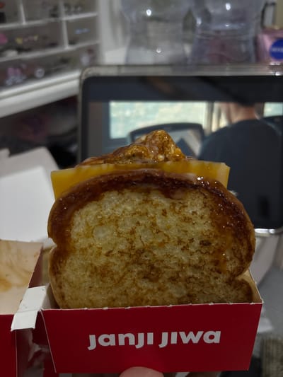 A close-up, vertical shot captures a breakfast sandwich, likely French toast with cheese, nestled within a red cardboard container. The sandwich is golden brown and appears slightly greasy, with a slice of melted yellow cheese peeking out from between two pieces of toast. The red container has white text printed vertically on its side, which reads "JANJI JIWA." The background is out of focus but suggests an indoor setting with a blurred reflection of what might be a tablet or screen, and a jewelry organizer with multiple compartments containing small items. The overall impression is that of a casual, possibly quick meal. No specific time of day or weather conditions are discernible. The location context of Tangerang, Indonesia, does not provide any visual cues within the image itself.