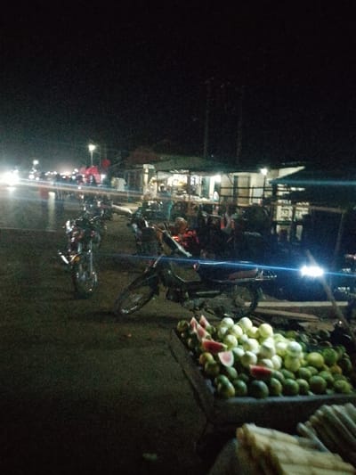 The image depicts a nighttime scene at a market stall in Serti, Nigeria. A variety of fruits, including watermelons and guavas, are displayed in a cart in the foreground. Several motorcycles are parked in the middle ground. In the background, a bustling market with stalls, lights, and moving vehicles suggests activity. The overall atmosphere is one of a lively nighttime marketplace.