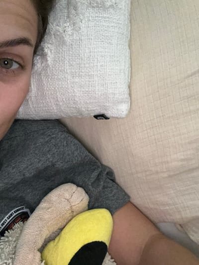 A person is partially visible, with one eye in focus. A white, textured pillow rests beside the person. A beige headboard or pillow is visible behind the white pillow. The person is wearing a gray garment, and a plush toy with yellow and beige fur is also present. The toy appears to have a sticker of some kind on it. The scene suggests a comfortable or relaxing environment, likely in a bed or resting space.