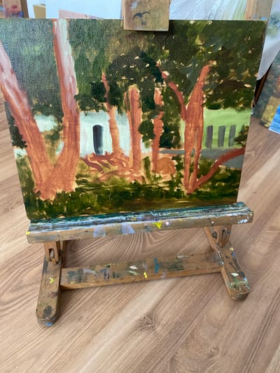 The image shows a painting on an easel, set on a wooden floor. The painting depicts a forest scene with tall, reddish-brown trees and lush green foliage. A small building with a dark doorway is visible in the background, surrounded by more trees and greenery. The easel is made of wood and shows signs of paint splatters, indicating it is used by an artist. The floor is made of light-colored wood with a visible grain pattern. The overall setting appears to be an indoor studio or workspace. No people are visible in the image. The location is not specified in the image.