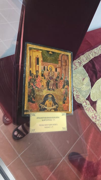 This image depicts an Eastern Orthodox icon displayed within a glass case in Bachkovo, Bulgaria. The icon, rendered in a traditional style with vibrant colors and gold accents, portrays a religious scene with multiple figures. A golden inscription plate below the icon provides information in both Cyrillic and Latin alphabets. The Cyrillic text reads "ЛИЧНА МОЛИТВЕНА ИКОНА НА ЕКЗАРХ АНТИМ I, "СВ. ПЕТДЕСЯТНИЦА", 1777г.", and the Latin text states "PERSONAL ICON OF EXARCH ANTIMUS I, "PENTECOST", 1777.". The icon is placed on a rich, dark red fabric with ornate gold embroidery. A dark red velvet-covered stand or box partially frames the icon. In the lower left corner, a person's foot wearing a sandal is visible, suggesting the viewer's presence. The flooring appears to be tiled. The overall setting indicates a museum or exhibition space likely within the Bachkovo Monastery, a significant religious and cultural landmark.