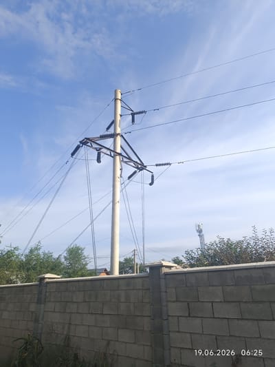 This image shows a view of a street in the city of Қырғауылды, Kazakhstan. In the foreground, there is a grey concrete block wall. Behind the wall, there are green trees and vegetation. A tall, light-colored utility pole with electrical wires is visible in the center of the image, extending into the blue sky with scattered white clouds. In the background, to the right, a cellular tower can be seen above the trees. The date and time stamp "19.06.2026 06:25" is visible in the lower right corner of the image. There are no people or animals present in the scene.