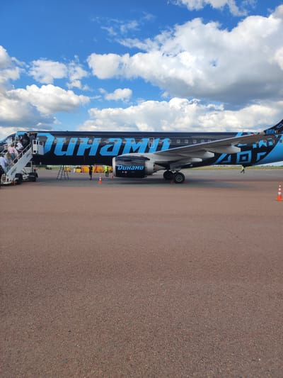 An Embraer E190 passenger jet, painted black with large, blue "DYNAMO" lettering and tribal-like blue patterns on the tail, is parked on an airport tarmac under a cloudy blue sky. A set of mobile stairs is attached to the aircraft's front-left door, with several passengers in casual attire boarding. The aircraft's engines are visible. In the background, a small bus or service vehicle with "DYNAMO" written on it is parked, and traffic cones are placed near the aircraft. The ground is a reddish-brown asphalt surface, common at airports. The location is the Пакалюбіцкі сельскі Савет in Belarus, likely at the Mogilev Airport, given the airline's branding.