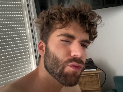 The image presents an indoor close-up portrait of an adult male. The subject, a Caucasian man with curly brown hair and a full beard, is positioned prominently in the foreground, winking his left eye and pouting his lips while looking directly towards the camera. A portion of his bare right shoulder and neck is visible.

To the left of the subject, a window is partially covered by horizontal louvered blinds, allowing strips of light to penetrate and create a pattern of parallel light and shadow. The background on the right features a light-colored wall. On this wall, a dark rectangular object, possibly a mounted television, is partially visible at the top. Below it, a wooden nightstand or small chest of drawers with carved panels displays a stack of dark books, a white remote control, and a black cable. A section of teal upholstered furniture is visible in the lower right corner. The scene is located within a private residence in Antibes, France.