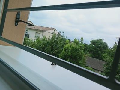 The image shows a view from a window in Blaye, France. The window has a grey metal frame with a latch mechanism attached to the wall. Outside the window, there is a building with a beige exterior and a red-tiled roof. Lush green trees and bushes fill the scene. A small building with a grey-tiled roof is visible amongst the trees. The sky is overcast.