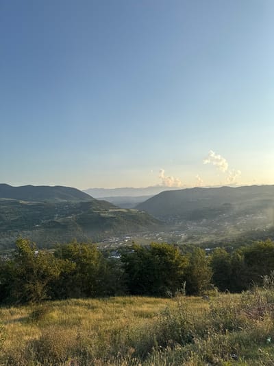 The image captures a panoramic view of a valley in the city of Tatil, Russia, bathed in the soft light of late afternoon or early morning. The foreground is dominated by a grassy hillside with patches of dry grass and weeds, hinting at a warm season. In the mid-ground, a dense line of trees forms a verdant border separating the hillside from the valley below.
The valley itself reveals a settlement with scattered buildings and rooftops, nestled amidst rolling, tree-covered hills. The landscape stretches into the distance, with successive layers of mountains fading into a hazy, atmospheric perspective. The sky above is a clear, pale blue, with a few wispy clouds tinged with the warm glow of the sun near the horizon. The overall impression is one of serene natural beauty and a peaceful rural setting.