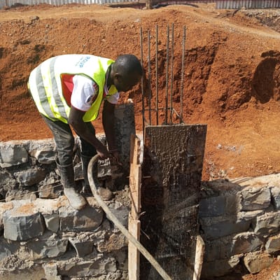 In Borabu Chitago ward, Kenya, a construction worker in a bright yellow safety vest, dark pants, and rubber boots uses a hose to apply material to a wooden form supporting vertical rebar. The form and rebar appear to be part of a foundation or supporting structure, set within a rock wall. Behind the worker, there is a mound of reddish-brown soil. The scene appears to be a construction site.