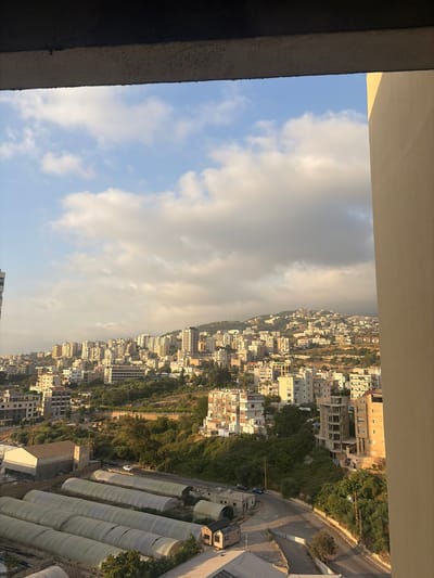 The image presents a panoramic view of a hillside city with a dense collection of buildings, suggestive of Wata Sillam, Lebanon. The city extends up a gentle slope, dotted with numerous residential and commercial structures. Many buildings are multi-story, with a variety of architectural styles, some appearing modern and others older. Lush green vegetation is interspersed throughout the urban landscape, particularly on the hillsides, indicating a natural environment integrated with the city. In the foreground, several long, tunnel-like structures, likely greenhouses or agricultural coverings, are visible. Roads wind through the scene, with a few cars present, suggesting active movement within the city. The sky is partly cloudy, with some large, diffused clouds obscuring parts of the sky and casting subtle shadows on the landscape, indicating it is likely late afternoon or early morning with the sun illuminating the buildings from the side. The overall impression is a bustling, lived-in urban environment situated within a verdant, hilly terrain.