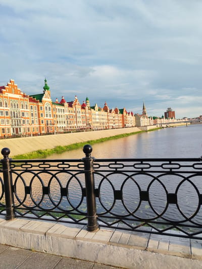 This media file depicts a picturesque waterside scene in Yoshkar-Ola, Russia. The primary subjects are a long, ornate row of buildings with colorful facades, predominantly orange and white with green and red accents on the roofs and spires. These buildings line a concrete embankment that separates them from a calm body of water. In the foreground, a black wrought-iron fence with decorative circular and square elements runs along a paved walkway.

The scene is bathed in the warm, soft light of either sunrise or sunset, suggesting it is either early morning or late evening. The sky is partly cloudy, with soft grey and white clouds scattered across a pale blue expanse. A few small, dark specks, possibly birds, are visible in the sky. The water of the river or canal reflects the sky and the warm light, creating a shimmering surface. Some lily pads or aquatic plants are dotted on the water's surface.

The architecture of the buildings is elaborate, with intricate window designs, balconies, and pointed turrets, evoking a European or historical aesthetic. Further down the river, a bridge can be seen, and beyond it, a taller, modern building stands out against the skyline. Small figures of people can be discerned walking along the embankment and seated at outdoor tables in front of some of the buildings, indicating a public space or promenade.

The overall mood of the image is tranquil and serene, showcasing the beauty of the urban landscape at a peaceful time of day. No visible text is present in the media file.