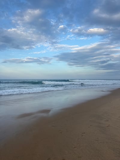 This media file captures a serene beach scene at what appears to be either sunrise or sunset, given the soft, diffused light and the color of the sky. The image is oriented vertically, presenting a panoramic view that divides the frame into distinct areas: the sky, the ocean, and the beach.
The left side of the image is dominated by a vast expanse of sky filled with dynamic clouds. These clouds are a mix of blues and whites, with some appearing wispy and others more solid, suggesting a dynamic atmosphere. The bright sunlight, though not directly visible, is filtering through these clouds, creating a luminous effect.
The middle section of the image depicts the ocean. Gentle waves are breaking along the shore, creating a foamy white line of surf. The water itself appears a tranquil blue-green. A few dark, indistinct shapes in the water suggest the presence of surfers or swimmers, engaged in activities like surfing or enjoying the water.
The right side of the image showcases a sandy beach. The sand is a light brown or beige color, with visible footprints, indicating recent human or animal activity. A shadow stretches across a portion of the sand, possibly cast by a person or an object not fully within the frame, adding depth to the scene.
The overall mood is peaceful and calming, evoking a sense of quietude and the natural beauty of a coastal environment. The location context, Arugam Bay, Sri Lanka, is known for its beautiful beaches and surfing spots, which aligns with the visual elements present in the image. There is no visible text in the media file.
