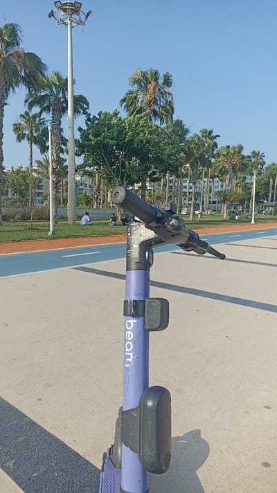 The image shows the handlebar and stem of a purple electric scooter, branded "Beam," in the foreground. The scooter is parked on a paved walkway. In the background, there is a park with palm trees, green grass, and people sitting on the grass. There is also a running track with blue and orange sections. A tall lamppost with multiple lights is visible on the left side of the image. Buildings can be seen in the distance. The sky is clear and blue. The scene appears to be in a public park or recreational area in Yenişehir, Turkey.