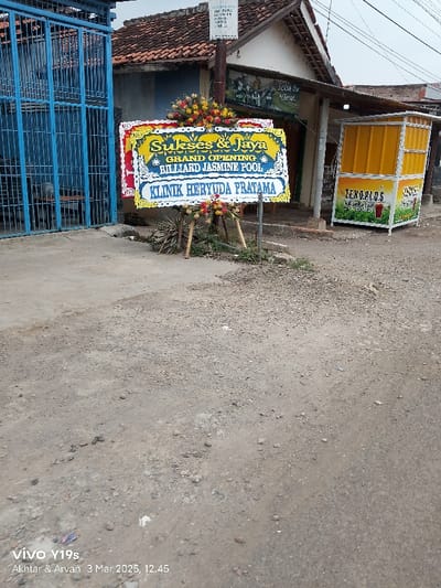 The image shows a street in Kecamatan Anjatan, Indonesia (latitude: -6.411, longitude: 107.961). There is a sign in front of a building that says "Sukses & Jaya Grand Opening Billiard Jasmine Pool Klinik Heryuda Pratama." The sign is decorated with flowers. There is a blue gate to the left of the sign and a yellow building with a "Lemarios" sign to the right of the sign. The ground is gravel. The image was taken with a mobile phone.