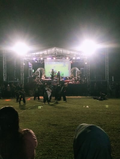 The image depicts a performance taking place in front of a stage in a field located in Kecamatan Sambirejo, Indonesia, at coordinates -7.5 latitude and 111.087 longitude. The stage is made of metal scaffolding and draped in a white tarpaulin. Several lights illuminate the stage, which features a band playing in front of a large screen displaying an image. The audience, many of whom are sitting on the ground, are watching the performance. There are a group of individuals in the foreground, some of whom are facing the camera, who appear to be performers or part of a procession. The field surrounding the stage is grassy, with a few pieces of litter scattered across it. The overall lighting of the scene is dim, except for the stage and the surrounding area. The sky is dark with no visible stars or clouds.