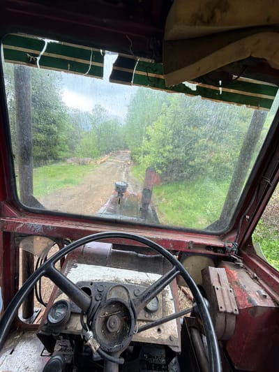 The image, taken from inside the rusted, worn cab of a vehicle in Рилски манастир, Bulgaria, features a view through the grimy windshield. The interior of the cab is decayed, with a black steering wheel dominating the right foreground and a damaged dashboard with gauges. Part of the interior is stained white. To the left of the windshield, remnants of a striped, possibly green and white, sun visor are visible. The view through the window reveals a dirt road leading into a lush green forest. There is a person standing some ways down the road, with another truck behind.