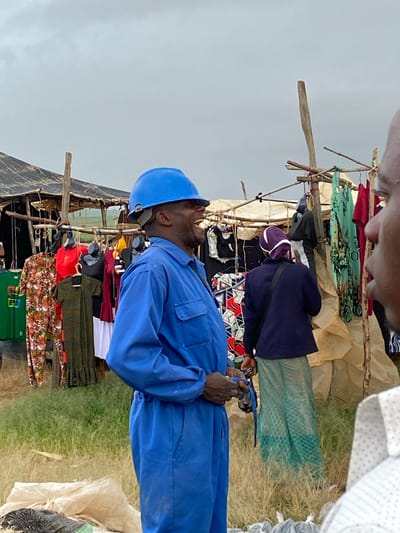 An outdoor market scene in Riyom, Nigeria, is captured under an overcast sky. In the foreground, a dark-skinned man wears a blue hard hat and a matching blue work suit, looking upwards and slightly to his right. He holds a small dark object, possibly a phone or a camera, in his hands.

Behind him and to his right, a market stall, crudely constructed from wooden poles and covered with what appears to be tarpaulin or fabric, displays various garments. These include patterned dresses and solid-colored tops hanging on hangers or draped over the wooden frame. Another person, seen from behind, stands near this stall, wearing a dark blue top and a white-and-purple cap. This individual appears to be examining the merchandise or tending to the stall. Further back and to the left, another similar market stall is visible, also laden with clothes for sale.

The ground beneath the stalls is covered with dry, sparse grass and dirt. In the upper part of the frame, another individual's head and shoulder are visible, looking down towards the market activity. The overall impression is of a bustling, informal clothing market, likely operating during the daytime under a cloudy sky. No discernible text is visible.