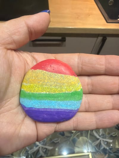 A human hand with light skin tone and a blue-painted thumb nail holds a small, oval-shaped rock. The rock is painted with five horizontal stripes in the following colors from top to bottom: red, yellow, green, light blue, and purple. The yellow stripe shows a textured or glittered finish. The background is out of focus, revealing a wooden surface in the upper right, a dark, indistinct object in the upper left, and various blurred shapes and reflections in the lower portion, suggesting an indoor setting.