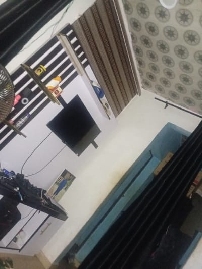 This image displays the interior of a domestic room in Ogbogbo, Nigeria, captured from an elevated and tilted perspective. The room features a white wall with black horizontal stripes, upon which two narrow wooden shelves are mounted. The top shelf contains a yellow container, an orange/yellow object, and a small package. The lower shelf holds a pink cup, a white ring-shaped object, and a clear plastic bottle. A black flat-screen television is mounted below the shelves on the striped wall, with visible cabling extending downwards. To the right, brown roller blinds adorned with a repeating geometric pattern are visible. The ceiling is covered with light-toned tiles, each featuring a central dark, star-like circular motif. In the lower right, a section of a dark blue wall or trim is present, alongside black vertical blinds. The lower left area includes a dark-topped, light-sided dresser or desk, cluttered with electronic components and wires. A framed picture, depicting a person in blue attire, leans against the wall on this surface. No individuals are present within the scene.