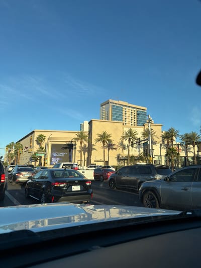 The image depicts a street scene in Las Vegas, Nevada. Several cars are stopped in traffic on a multi-lane road. Tall buildings line the street, including a tan-colored building with palm trees and a large electronic billboard advertising "Magic Mike Live". The sky is clear and blue. Traffic lights and street signs are visible at intersections.