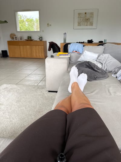The image shows a person lying on a light gray sofa. The person is wearing brown shorts and white socks, with their legs crossed at the ankles. The sofa is cluttered with pillows, blankets, and clothing. In the background, there is a cabinet, a window, and framed artwork on the wall. The room has white walls and a light-colored tile floor. The location is potentially Veauchette, France, based on the presence of typical European architectural features and interior design. No specific elements definitively confirm the location, but it remains a possibility.