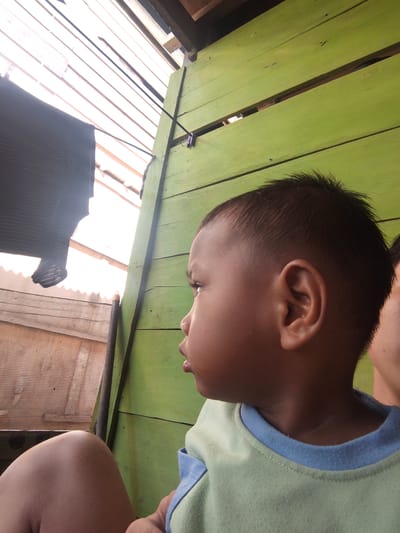 An outdoor, eye-level, medium shot captures a close-up profile of a young child, facing right. The child's skin tone is brown, and their hair is dark and short, styled with a high fade. They are wearing a light green t-shirt with a blue collar. The background is a bright, wooden structure painted green, with horizontal planks. To the left, a dark, striped fabric hangs, possibly laundry, against a bright, white background that appears to be a corrugated roof or wall. A dark, vertical pole is visible in the left portion of the frame, suggesting a structural element. The overall scene suggests a residential or outdoor area in Pompanua, Indonesia, possibly a home or yard, characterized by simple construction and natural light.