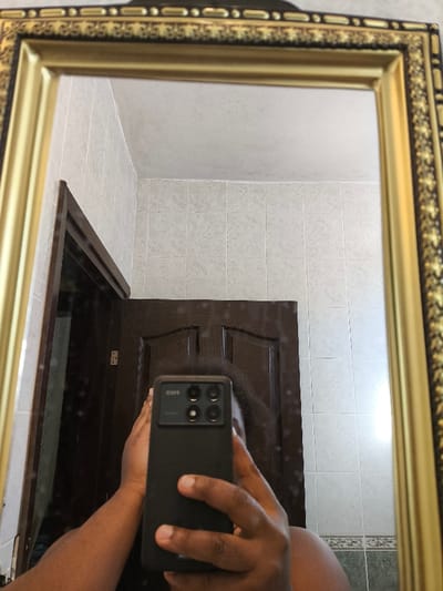 A selfie is taken in a framed mirror in a bathroom. Light-colored square tiles line the walls. A partially visible, dark wooden door is in the background, slightly ajar. The person taking the photo is holding a black smartphone. Their face is obscured by the phone and their hands, with a glimpse of dark hair visible. The mirror has an ornate gold and dark-colored frame.