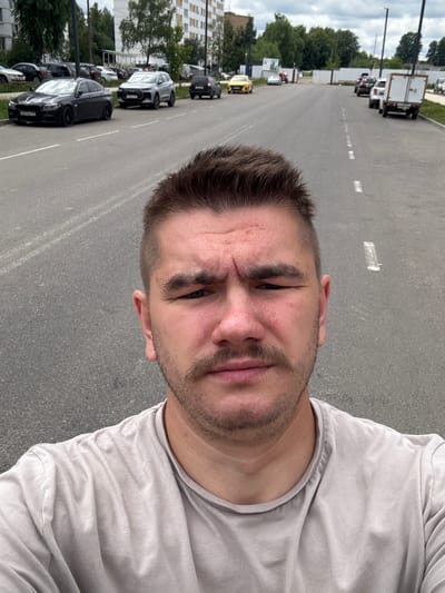 This is a vertical, selfie-style photo of a young man in Shcherbinka, Russia. The man is in the foreground, his face and upper torso filling the right side of the frame. He has short, dark hair, a beard, and a light-colored t-shirt. His expression is neutral. The background shows a street with parked cars and a building with many windows.

The photo appears to have been taken outdoors during the daytime. The lighting suggests it might be overcast or late afternoon. The environment seems urban, with a paved road and sidewalks, typical of a residential or commercial area. The presence of multiple cars suggests a parking lot or a street with ample parking. The building in the background looks like an apartment complex.

No discernible text is visible in the image, other than possibly license plates on the cars which are too blurry to read. The man's posture and the framing of the shot suggest he is taking a casual picture of himself in his surroundings.