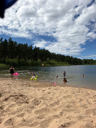 This is a vertically oriented, high-angle, medium shot of a sandy beach and a lake on a sunny day. The beach is in the foreground, with a person in a black swimsuit standing in the shallow water on the right side of the image. A pink inflatable object is near their feet. Several other people are in the water further out, some swimming, some standing. A person in a yellow inflatable float is visible in the middle of the lake. The lake stretches into the distance, bordered by a dense forest on the left side. The sky above is blue with scattered white clouds. The overall impression is one of a casual, recreational day at the lake. The time of day appears to be daytime, given the bright sunlight. The weather seems pleasant, with a clear blue sky and clouds, suggesting a warm day suitable for swimming. The location appears to be a natural setting, likely a lake or a reservoir with a sandy shore, surrounded by coniferous trees. There is no visible text in the image.