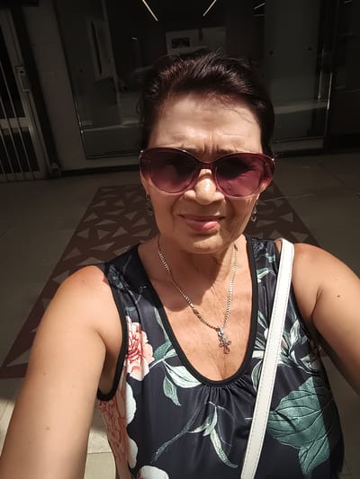 A middle-aged woman with dark hair and sunglasses is taking a selfie. She is wearing a sleeveless dark floral print top and a silver chain necklace with a cross pendant. A white strap from a bag is visible across her shoulder. She is standing outdoors on a tiled walkway in what appears to be a modern urban setting. The background includes a building facade with glass doors and vertical blinds, and the ground features a patterned tile design casting a shadow. The lighting suggests it is daytime, possibly late afternoon, given the strong shadows. There is no specific information about the location in Livorno, Italy, other than the context provided by the prompt.