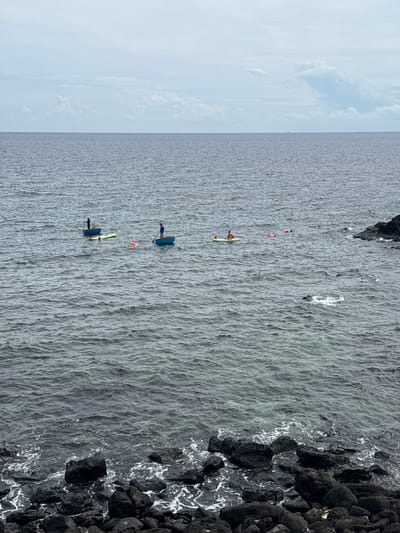 The image shows a body of water with several people engaged in water activities. In the distance, there is a hazy horizon with a cloudy sky. The foreground is dominated by dark, wet rocks along the shoreline.
Closer to the shore, there are three small blue boats with individuals in them. One boat has a person standing, holding a paddle. Another boat has a person in it, appearing to be steering. A third boat is partially visible.
Further out in the water, there are at least three people on paddleboards or similar flotation devices. One person is on a green paddleboard, and another is on a lighter colored one. There are also orange buoys or life vests scattered in the water. The water itself is choppy with small waves. The overall scene suggests a recreational or possibly work-related activity on the sea near the shore. Based on the context of Lý Sơn Special Administrative Region, Vietnam, this could be a fishing or water sports scenario.