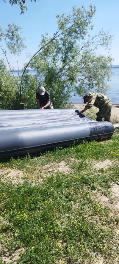Two men are working on an inflatable boat on the shore of a body of water. The man on the left is wearing a dark striped t-shirt and a baseball cap, and is bent over the boat. The man on the right is wearing camouflage clothing and is also bent over the boat, appearing to be adjusting it. The boat is dark gray and is partially deflated or being prepared for inflation. The environment is grassy with scattered yellow wildflowers. Behind the boat and men, there are trees and the water of a lake or river. The sky is clear and blue. The scene suggests an outdoor recreational activity.