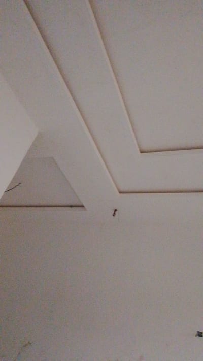 The image shows an interior view of a room in Enugwu-Ukwu, Nigeria. The focus is on the ceiling, which features a recessed geometric design. The ceiling and walls are painted in a similar shade of off-white. There are two visible recessed portions in the ceiling. One has a triangular shape and the other has a larger geometric design with intersecting rectangular elements.