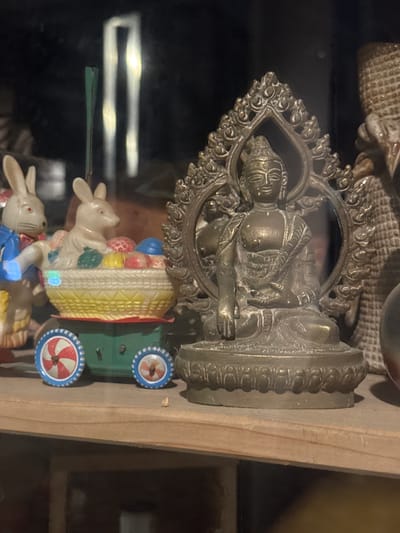 A silver Buddha statue with an ornate halo sits atop a green and white toy cart pulled by two white rabbits. The scene is indoors, likely a shelf or display case, with a wooden panel visible on the right. The location is Givatayim, Israel.
